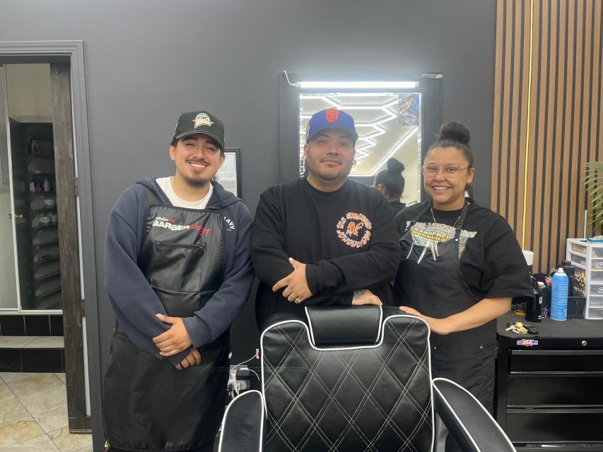 Three barbers stand behind a black salon chair, smiling at the camera in a modern barbershop with dark walls and wood accents.