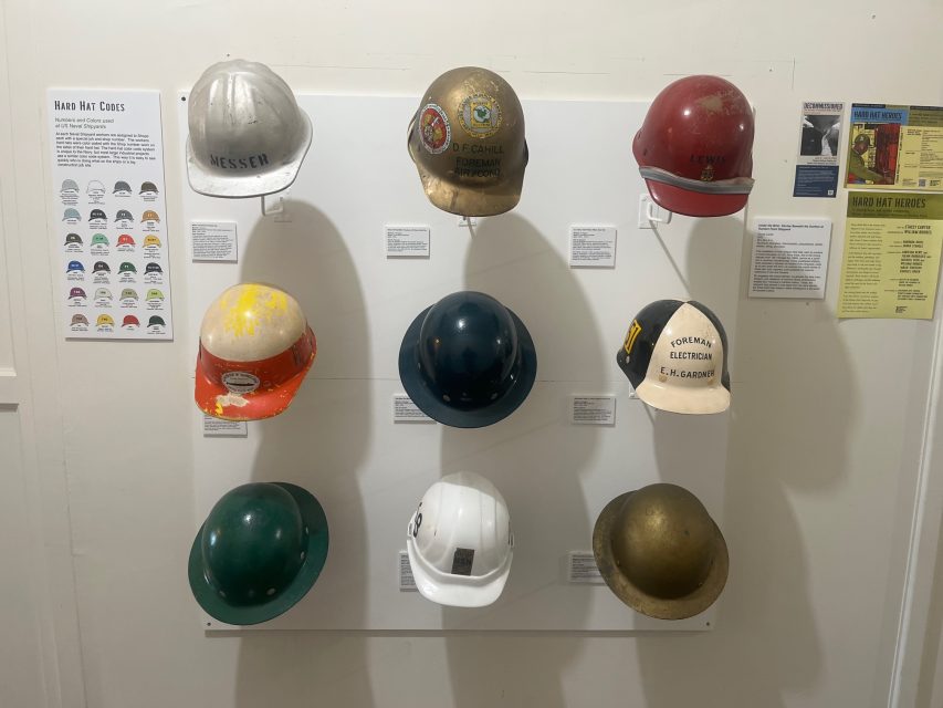A display of nine vintage hard hats in three rows, each with unique colors, logos, and labels, mounted on a wall with informational posters beside them.