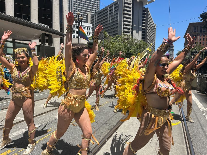 A group of dancers in gold costumes with yellow feathered wings parade down a city street with their arms raised, celebrating pride amid the towering buildings.