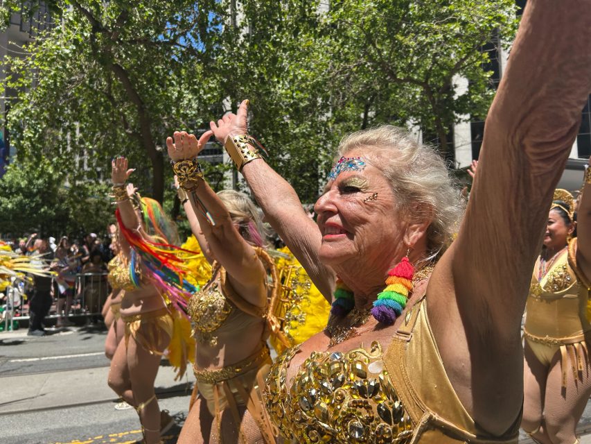 A group of older women in gold costumes and rainbow accessories march in a pride parade with their arms raised, surrounded by trees and spectators.