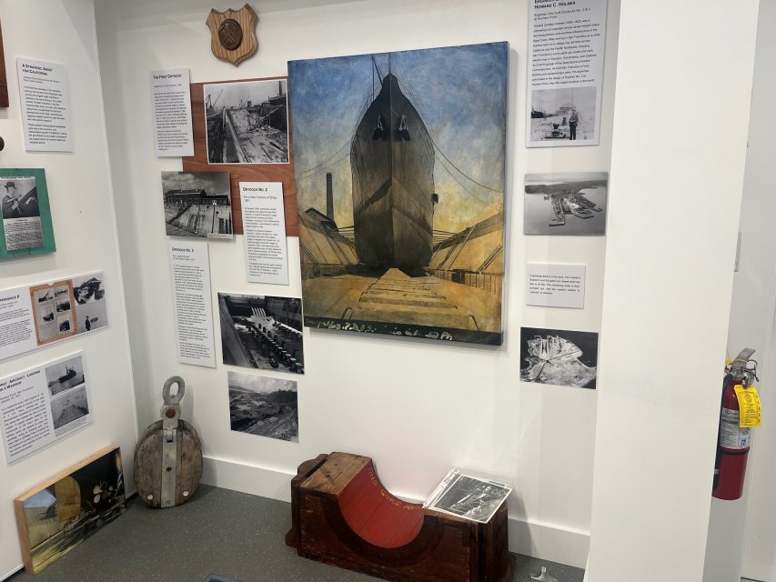 A museum exhibit wall displaying shipbuilding photos, informational plaques, a ship painting, and maritime artifacts, including a pulley, a red wooden piece, and a fire extinguisher.