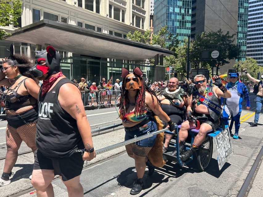 A group of people in colorful pride costumes and pet gear participate in a lively parade, some pulling a cart with others riding, on a city street lined with tall buildings.