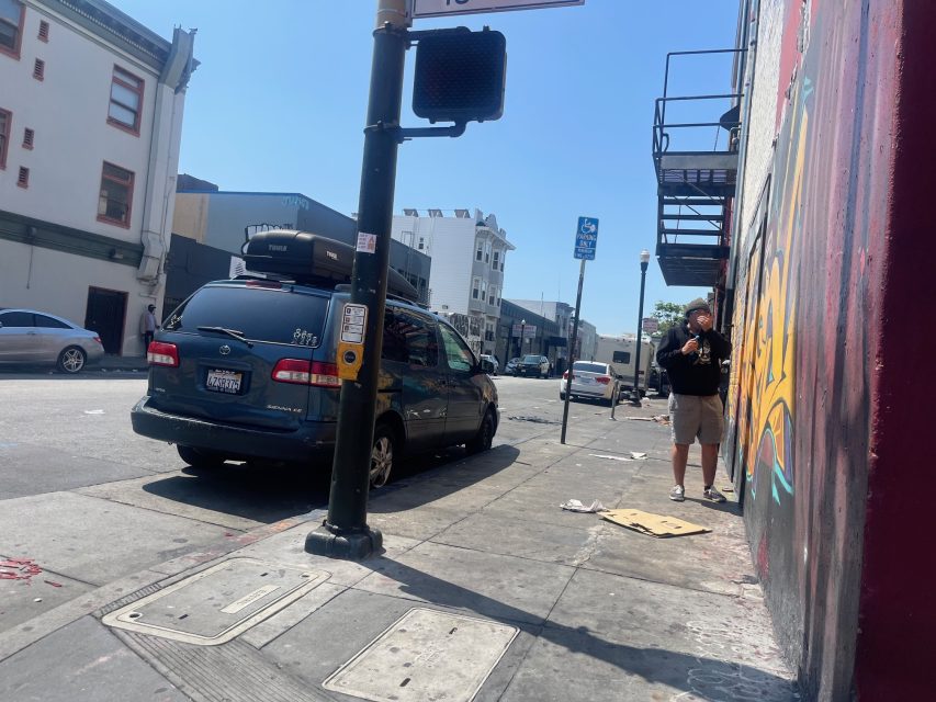 A man stands on a city sidewalk beside a mural, near a van parked by a curb under a clear blue sky.