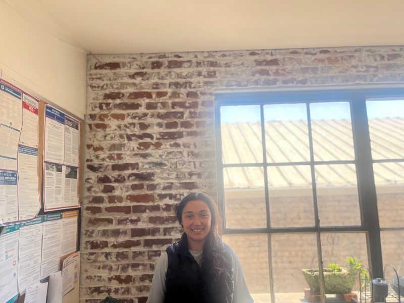 A person stands in an office with exposed brick walls, a large window letting in sunlight, and several notices posted on a bulletin board.