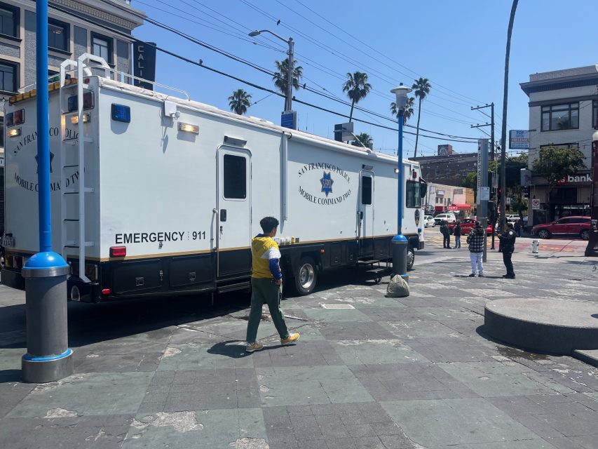 A large San Francisco Police Department mobile command vehicle is parked in a city plaza as pedestrians walk nearby on a sunny day.