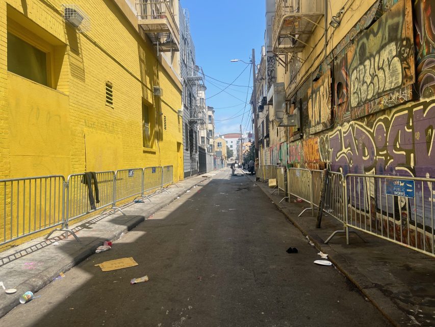 A narrow urban alley with yellow walls on the left, graffiti on the right, metal barricades, and scattered trash along the pavement under a clear sky.