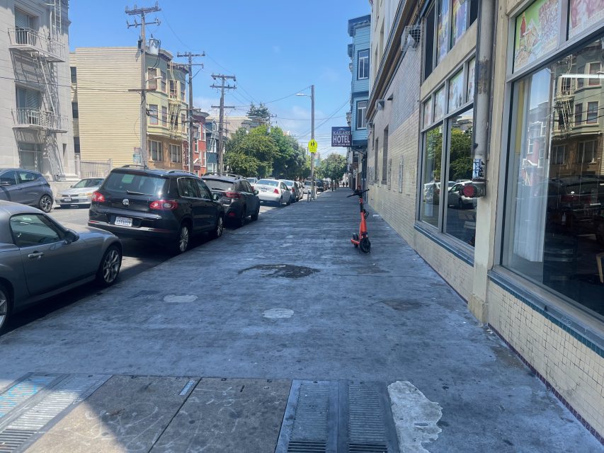 A city sidewalk with parked cars along the street, a scooter standing upright near a building, and apartment buildings in the background on a clear day.