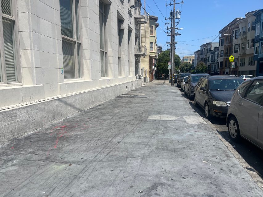 A city sidewalk runs alongside a light-colored building with parked cars on the street and apartment buildings in the background under a clear sky.