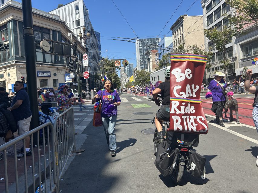 Person rides a bike with a sign reading "Be Gay Ride Bikes" at a pride city parade, while others walk and spectate in the sunny urban setting.