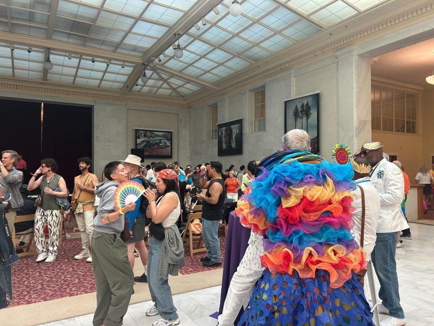 People gather in a large hall for a pride celebration, some wearing colorful outfits and accessories, while others converse and take photos. The space has high ceilings and framed artwork on the walls.
