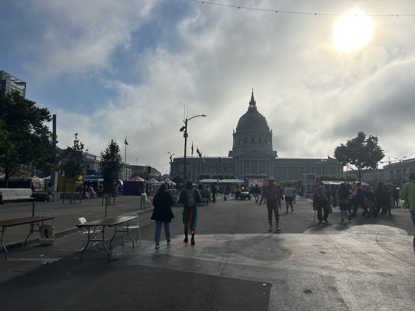 People walk in an open plaza with tables and tents, many displaying pride colors, facing a large domed government building under a partly cloudy sky.