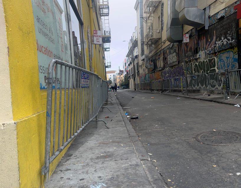A narrow urban alley with graffiti-covered walls, metal barricades, scattered litter, and a few people in the distance under an overcast sky.
