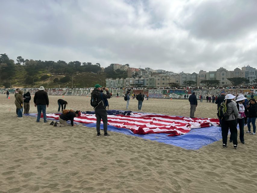 People gather on a sandy beach around a large American flag spread out on the ground in protest, with houses and trees visible in the background under a cloudy sky.