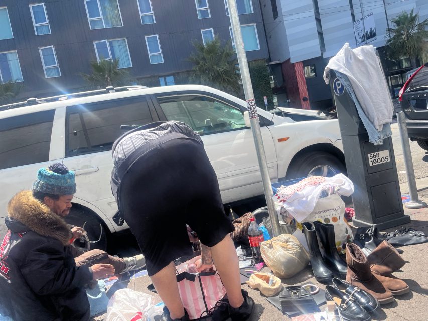 Two people sort through various belongings and clothing laid out on a sidewalk next to a white vehicle and a parking meter in an urban area.