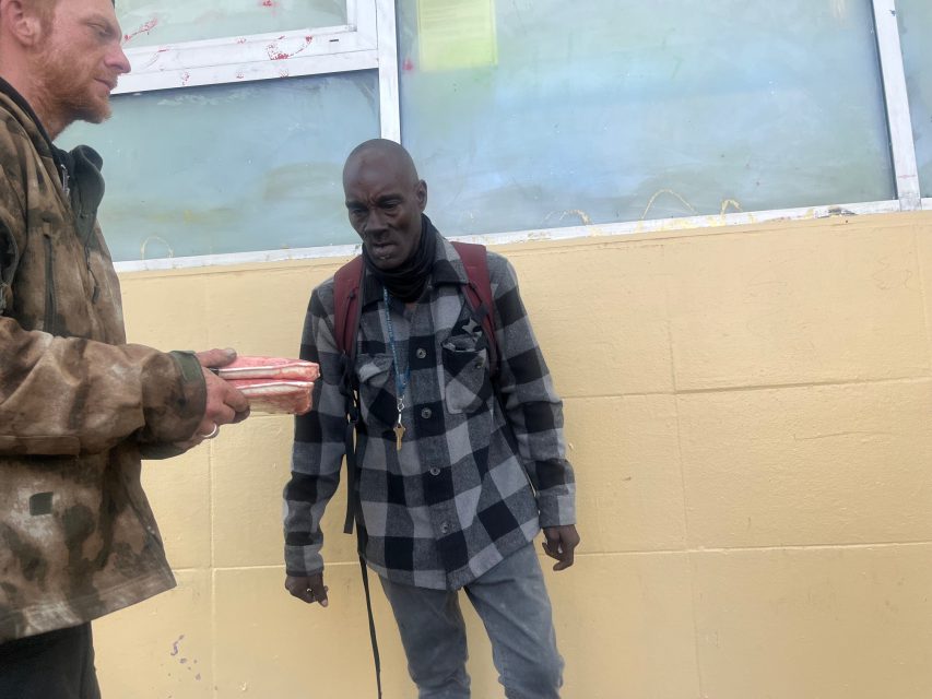 A man in a plaid jacket and red backpack stands outside against a yellow wall while another person offers him a package of meat.