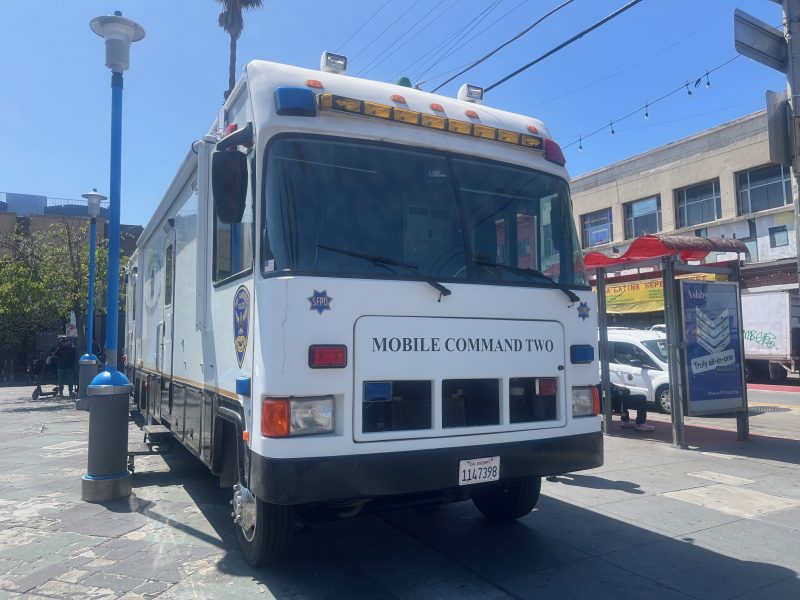 A white police mobile command vehicle labeled "Mobile Command Two" is parked on a city street near a bus stop under a clear blue sky.