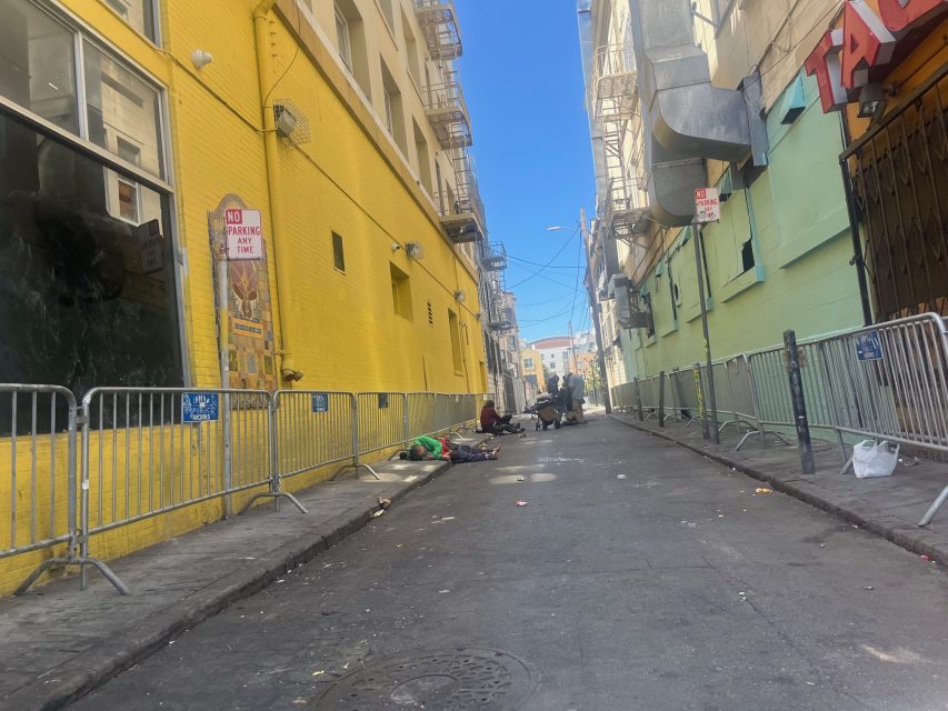 A narrow alley with yellow and green walls, metal barriers, and scattered debris; several people are lying on the ground along the sides of the alley.