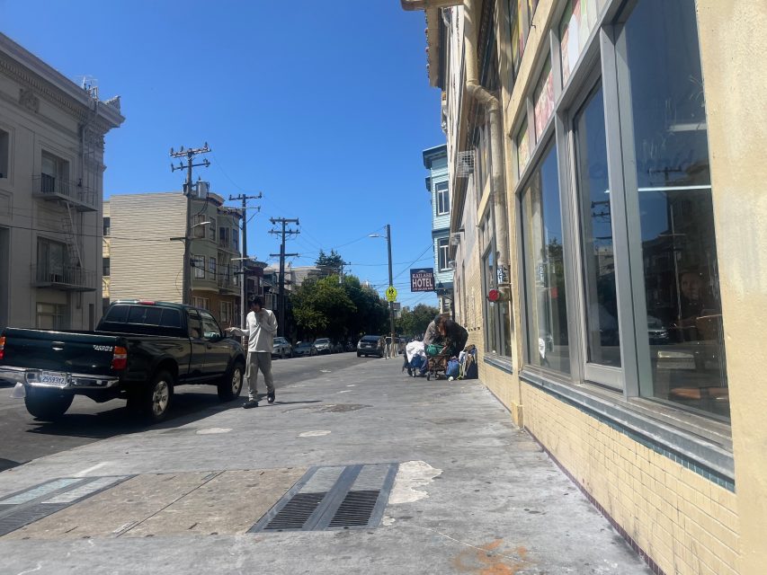 A city sidewalk with a few people gathered near a building, a parked truck, and clear blue sky above.