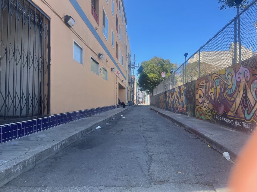 A narrow urban alley with graffiti on the right wall, a beige building on the left, scattered litter, and a few trees visible in the distance under a clear blue sky.
