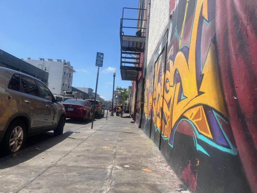 A city sidewalk with parked cars, a streetlamp, and a colorful graffiti mural covering the wall on the right side. Blue sky is visible above.