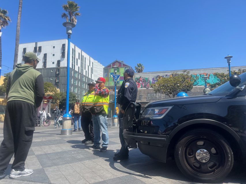 Two men in safety vests and a police officer stand next to a police car, talking to a man in a green hoodie on a sunny city street with palm trees and graffiti in the background.