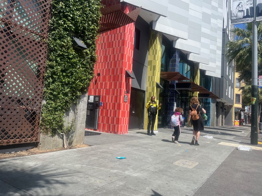 Three people walk on a sunny city sidewalk in front of a modern building with red, green, and blue columns and leafy plants on the wall.