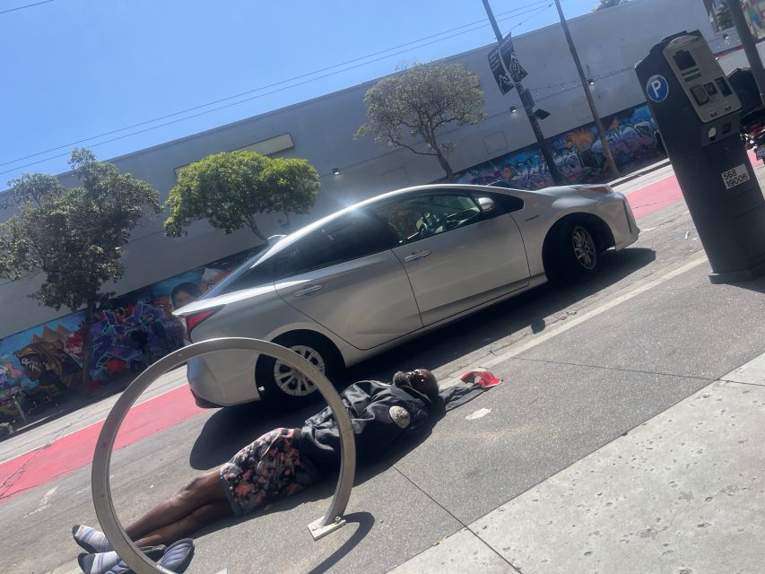 A person lies on the sidewalk near a bicycle rack, with a parked silver car and a parking meter nearby; street art is visible on the wall in the background.