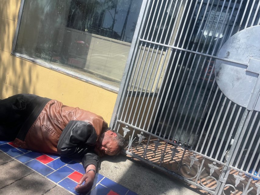 A man in a brown and black jacket lies on a tiled sidewalk beside a yellow wall and a metal gate in daylight.