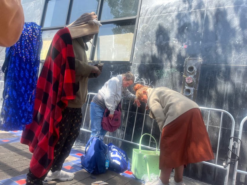 Three people stand and lean over bags on a sidewalk beside a tiled curb and a black wall with graffiti; one is covered in a red plaid blanket.