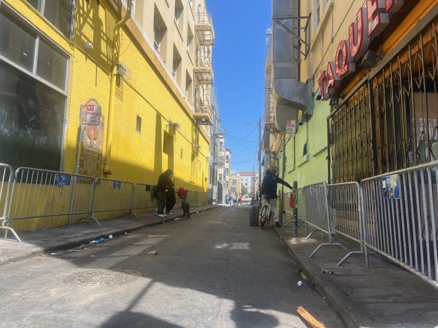 A narrow city alley with yellow and green buildings, metal barricades, and people standing and walking in the sunlight.
