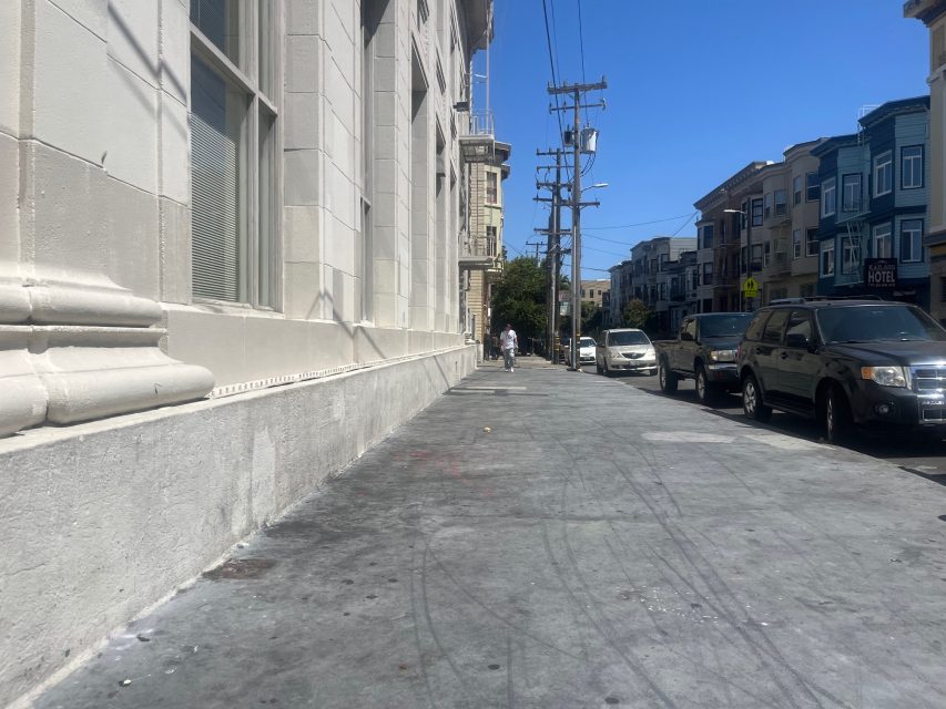 A city sidewalk runs alongside a beige building with parked cars and utility poles lining the street on a clear, sunny day.