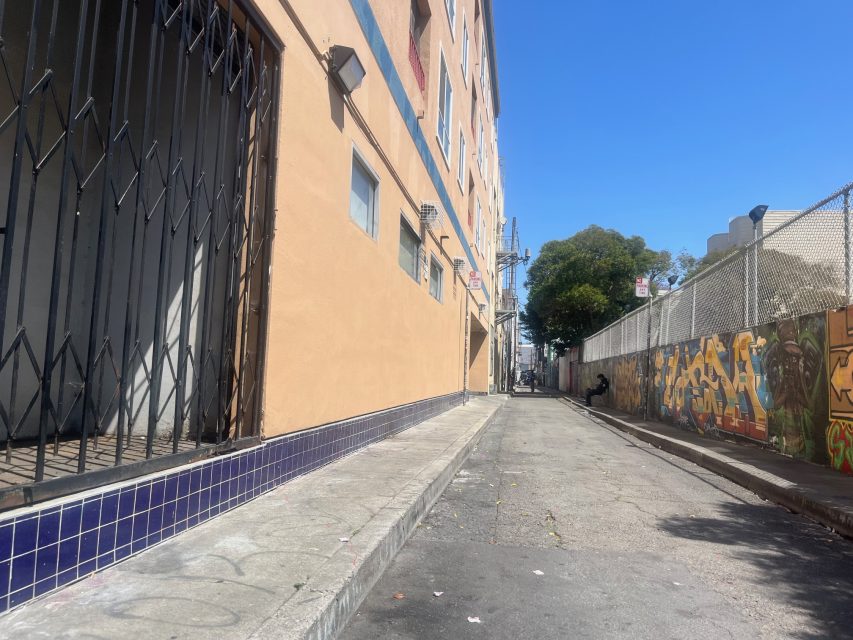 A narrow urban alleyway with a beige building on the left, graffiti-covered wall on the right, and a clear blue sky overhead.