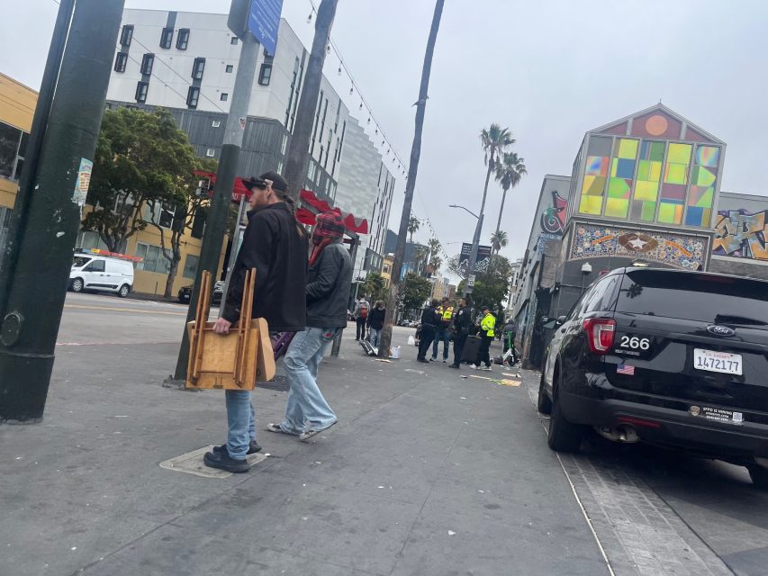 A man carries stacked wooden chairs on a city sidewalk; police and several people are gathered in the distance near parked cars and buildings adorned with mural art.