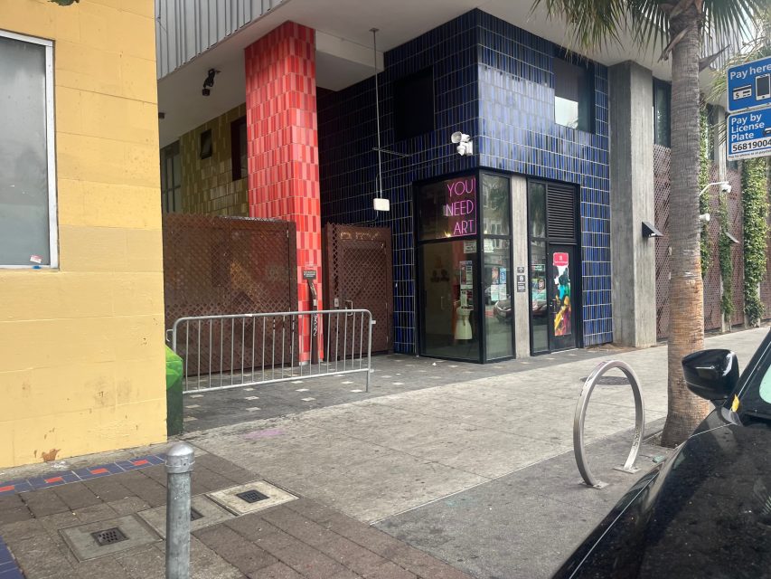 Street view of a building with colorful yellow, red, and blue tile walls, a "YOU NEED ART" sign in the window, and a metal barricade on the sidewalk—celebrating creativity in urban art spaces.