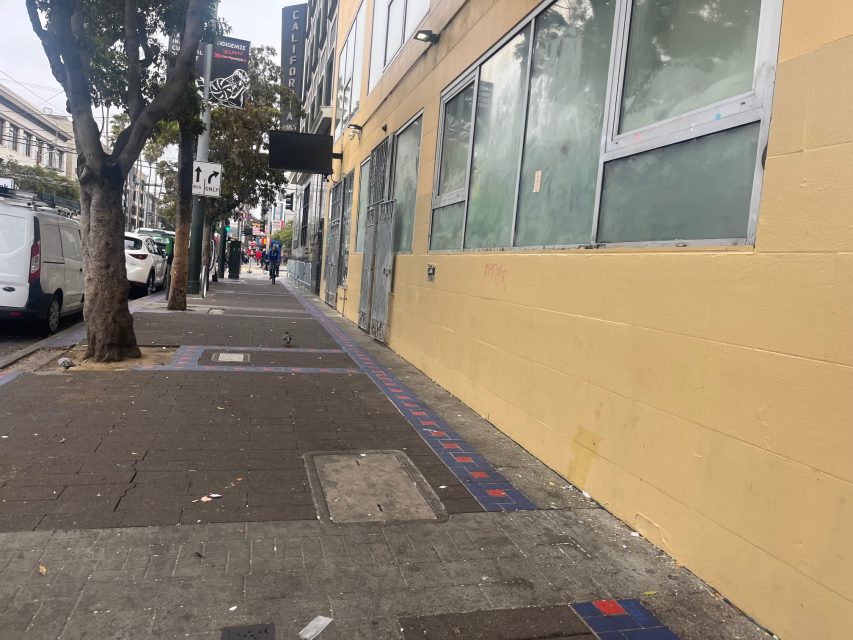 A city sidewalk with trees, parked cars, and a yellow building with covered windows creates an artful scene; a few people and pigeons are visible in the distance.