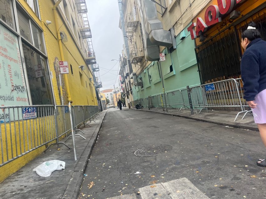 A narrow urban alleyway lined with yellow and green buildings, metal barricades along both sides, and a woman standing on the right in foreground, evokes the feel of urban art and city life.