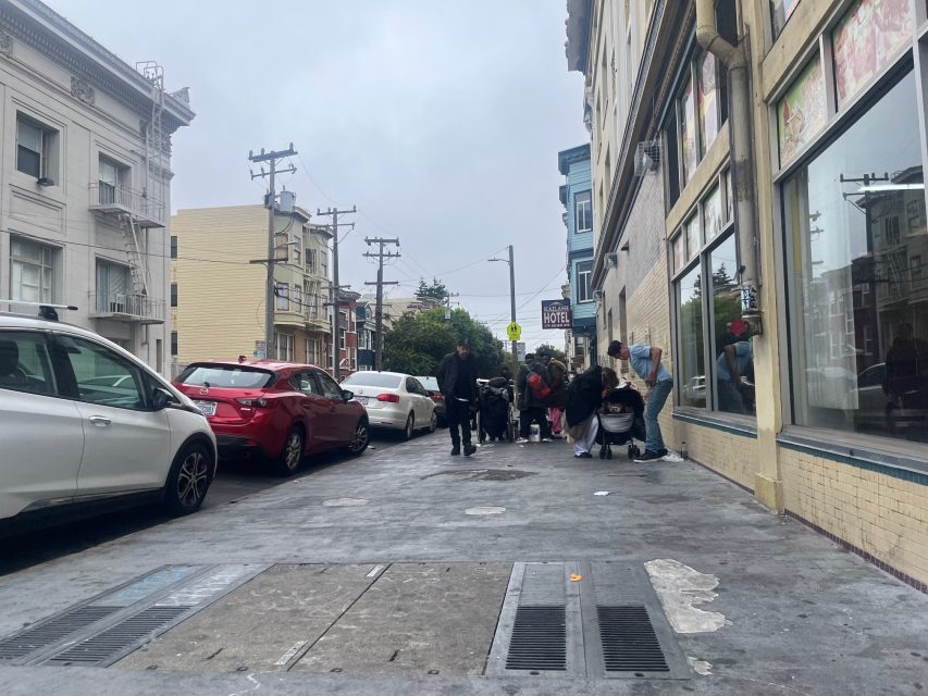A group of people gather on a city sidewalk near parked cars and buildings on a cloudy day, creating a scene reminiscent of urban art.