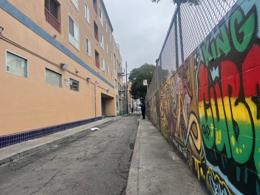 A narrow urban alley with cracked pavement, a beige apartment building on the left, and a graffiti-covered wall showcasing vibrant street art on the right, under a cloudy sky.