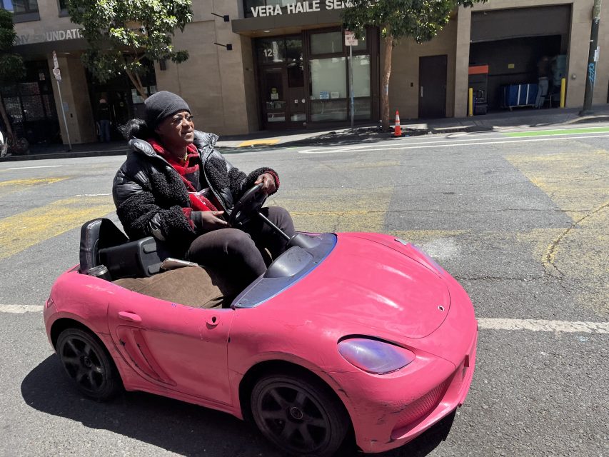 A person wearing a black beanie and jacket drives a small pink toy car on a city street in daylight.