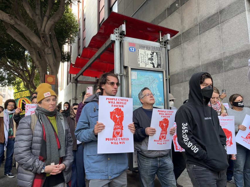 A group of people stand outside a building holding signs that read "People United Will Defeat Fascism" and "People United Will Win" during a protest.