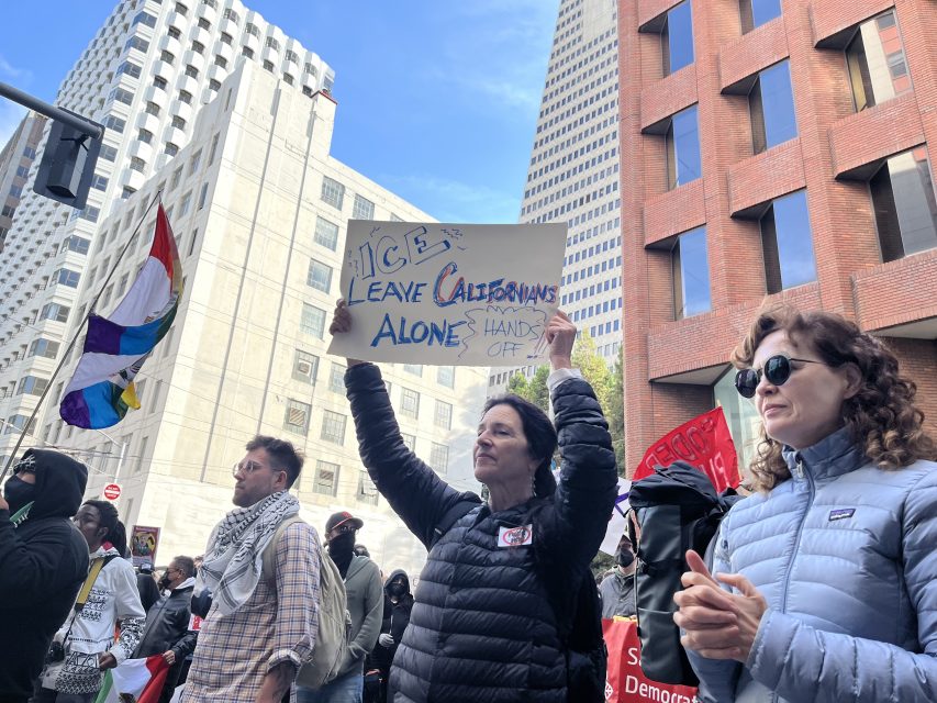 A person holds a sign reading "ICE: Leave Californians Alone! Hands Off" at a protest, surrounded by other demonstrators in a city with tall buildings in the background.