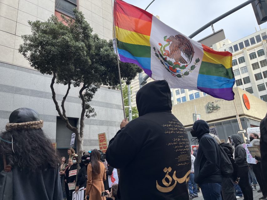 Person holds a rainbow flag with the Mexican coat of arms at a city street protest; other people stand nearby holding signs.