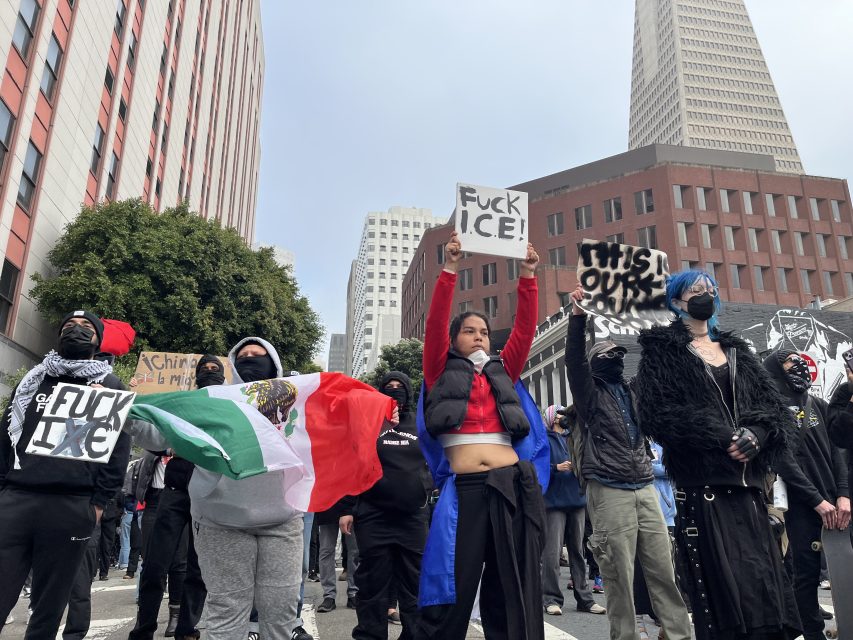 A group of protesters stand in a city street holding signs and a Mexican flag; some wear masks and dark clothing. Tall buildings are visible in the background.