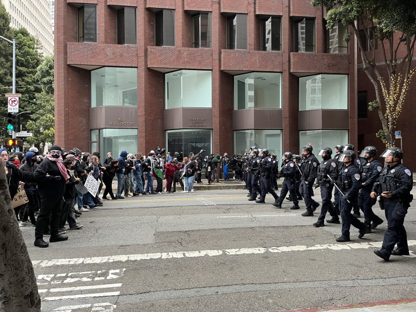 A line of police officers in riot gear face a crowd of protesters holding signs on a city street in front of a brick building.