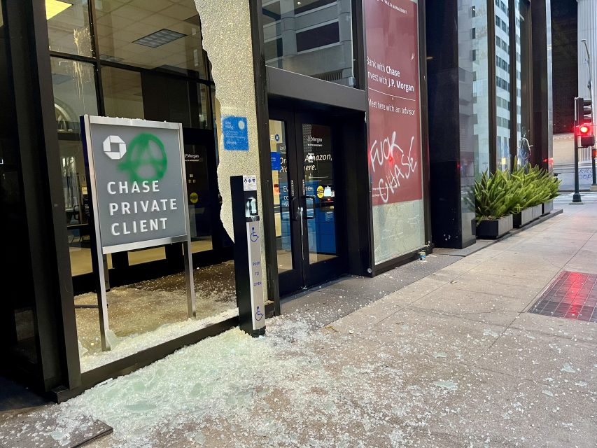 Broken glass and shattered windows at the entrance of a Chase Private Client bank branch; graffiti is spray-painted on the building’s exterior wall.