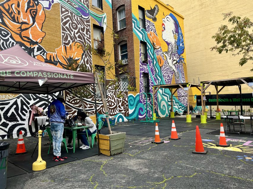 Outdoor urban space with colorful murals, people sitting under a canopy labeled "BE COMPASSIONATE," traffic cones, and planters on a paved lot next to a brick building.