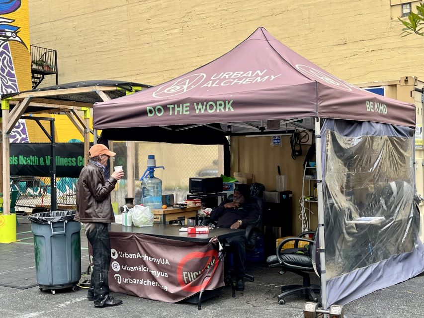 A man stands outside an Urban Alchemy tent, where another person sits at a desk beneath the canopy in an urban outdoor setting.