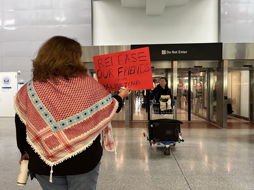 A person holding a red sign that reads "Release our friends from Palestine" stands in an airport near the "Do Not Enter" doors.