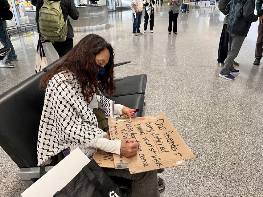 A Palestinian person wearing a mask sits on an airport chair, writing on a cardboard sign with a marker. Other travelers are visible in the background.