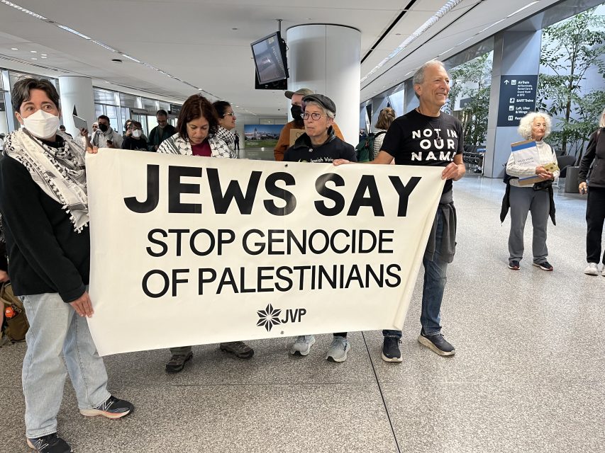 A group of people in an indoor public space hold a banner reading, "JEWS SAY STOP GENOCIDE OF PALESTINIANS JVP," showing solidarity with the Palestinian people.